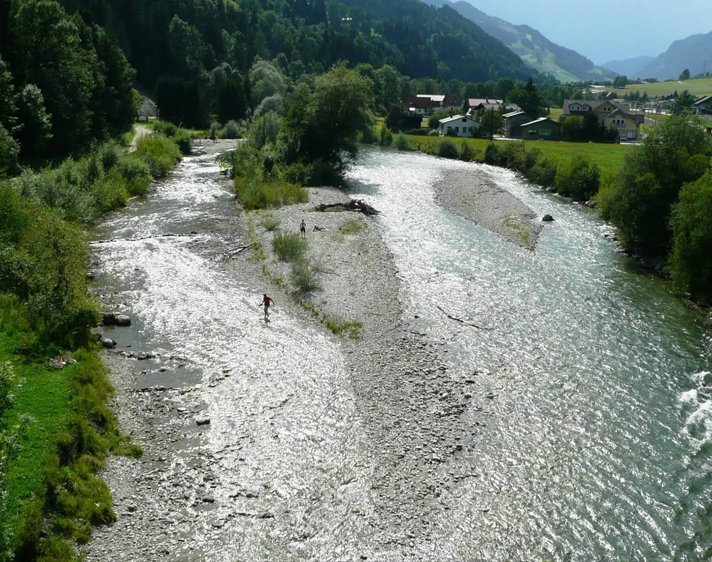 A river meanders through a rural setting, with a small island in its midst. People stroll along the gravel bed, surrounded by lush trees and traditional houses in the background.